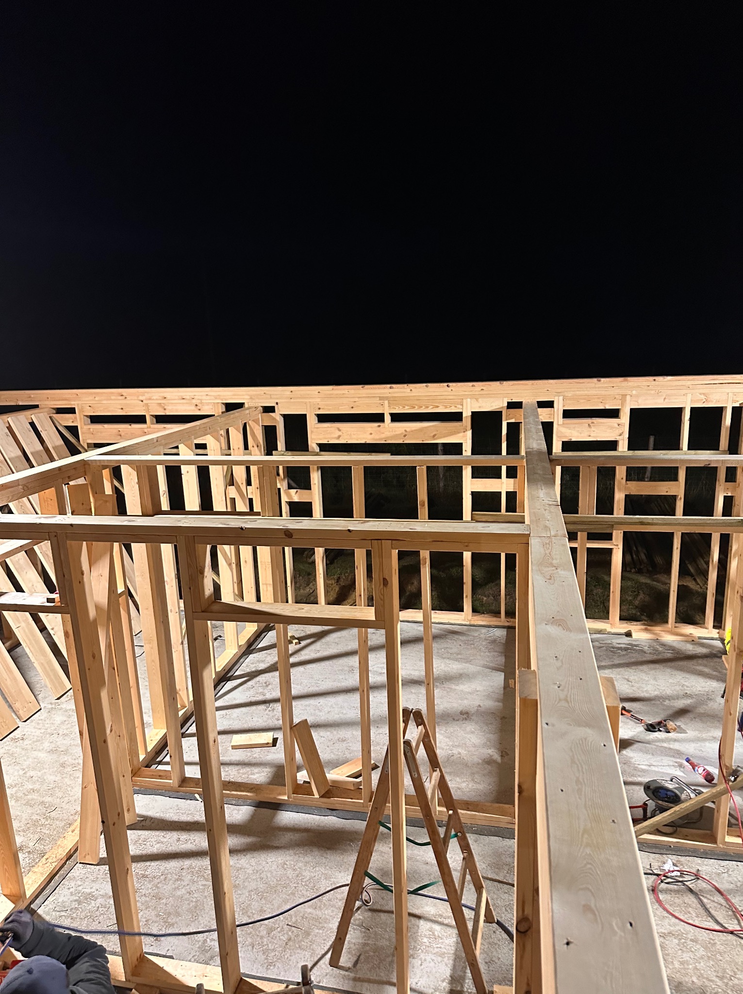 Wznoszenie drewnianej konstrukcji szkieletowej budynku nocą, widoczne narzędzia i drabina, surowy stan, ciemne tło.