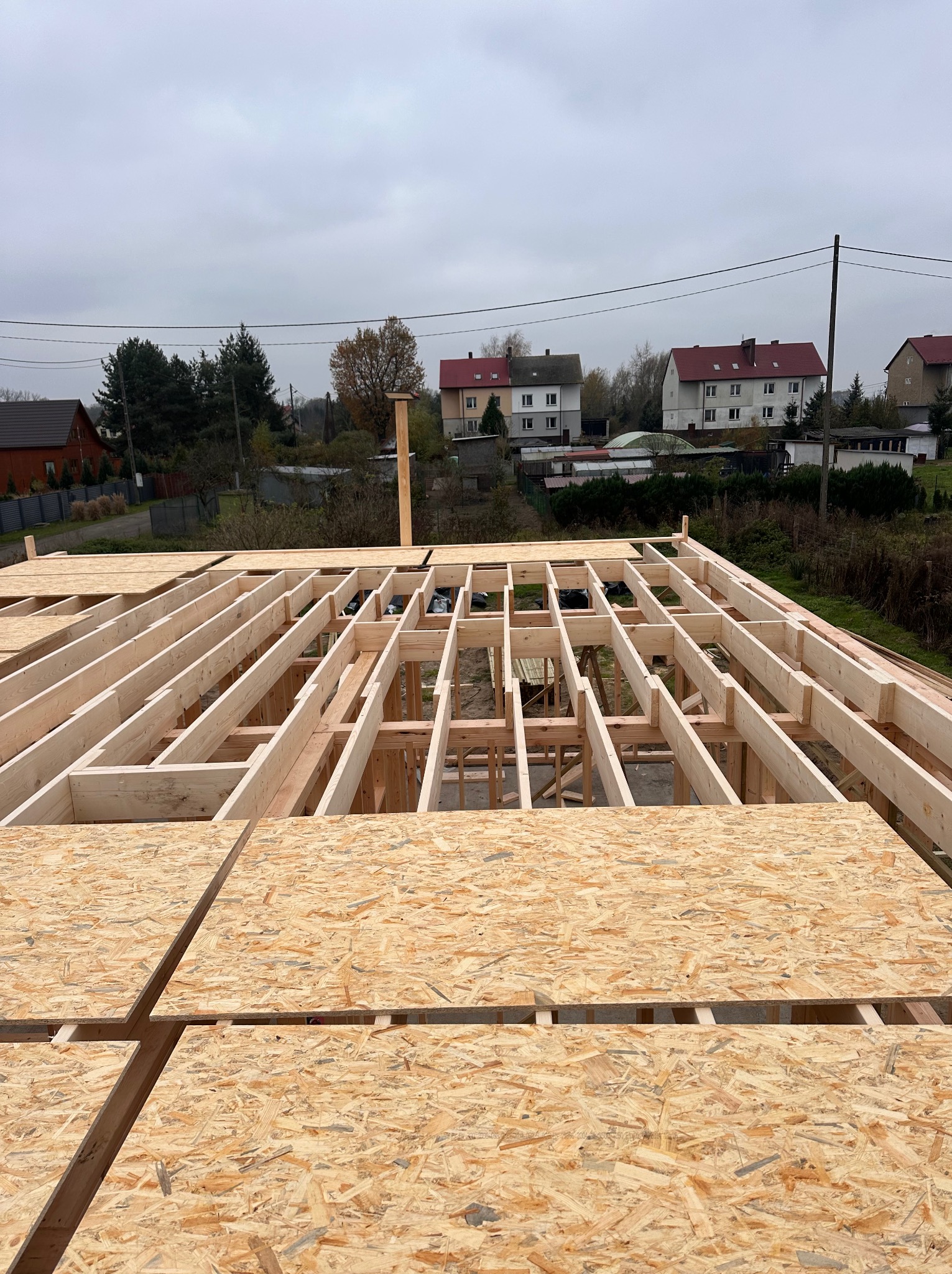 Konstrukcja drewniana dachu w trakcie budowy, widoczne płyty OSB i elementy szkieletu, w tle zabudowa jednorodzinna w pochmurny dzień.