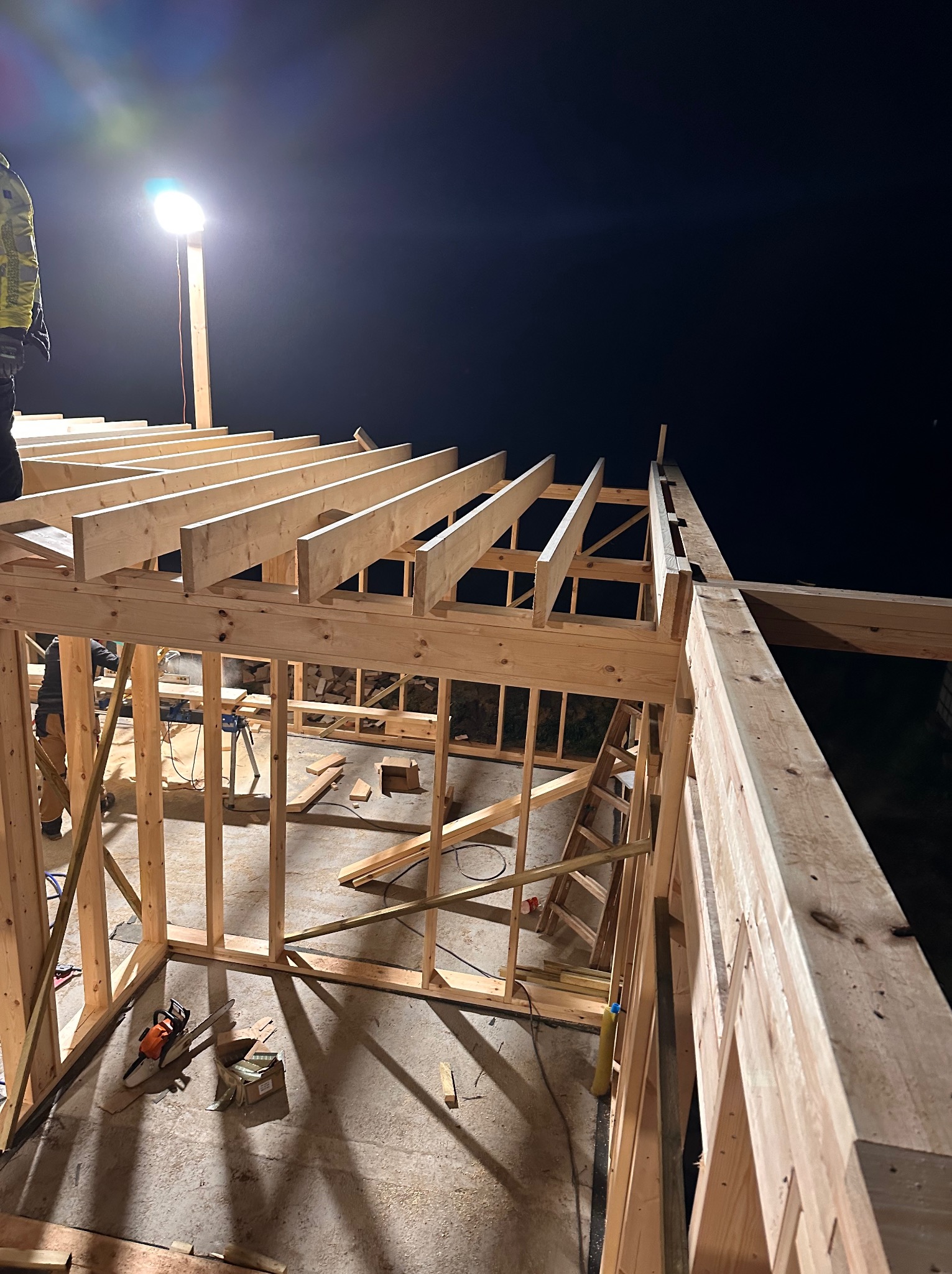 Konstrukcja drewniana domu w trakcie budowy nocą, oświetlona lampą budowlaną, widoczne elementy szkieletu i więźby dachowej, piła łańcuchowa leżąca na betonowej posadzce.