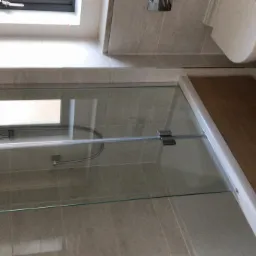 Nowoczesna kabina prysznicowa z szklanymi drzwiami, widoczny fragment toalety i okno dachowe, szare płytki na ścianach, drewnopodobny brodzik.