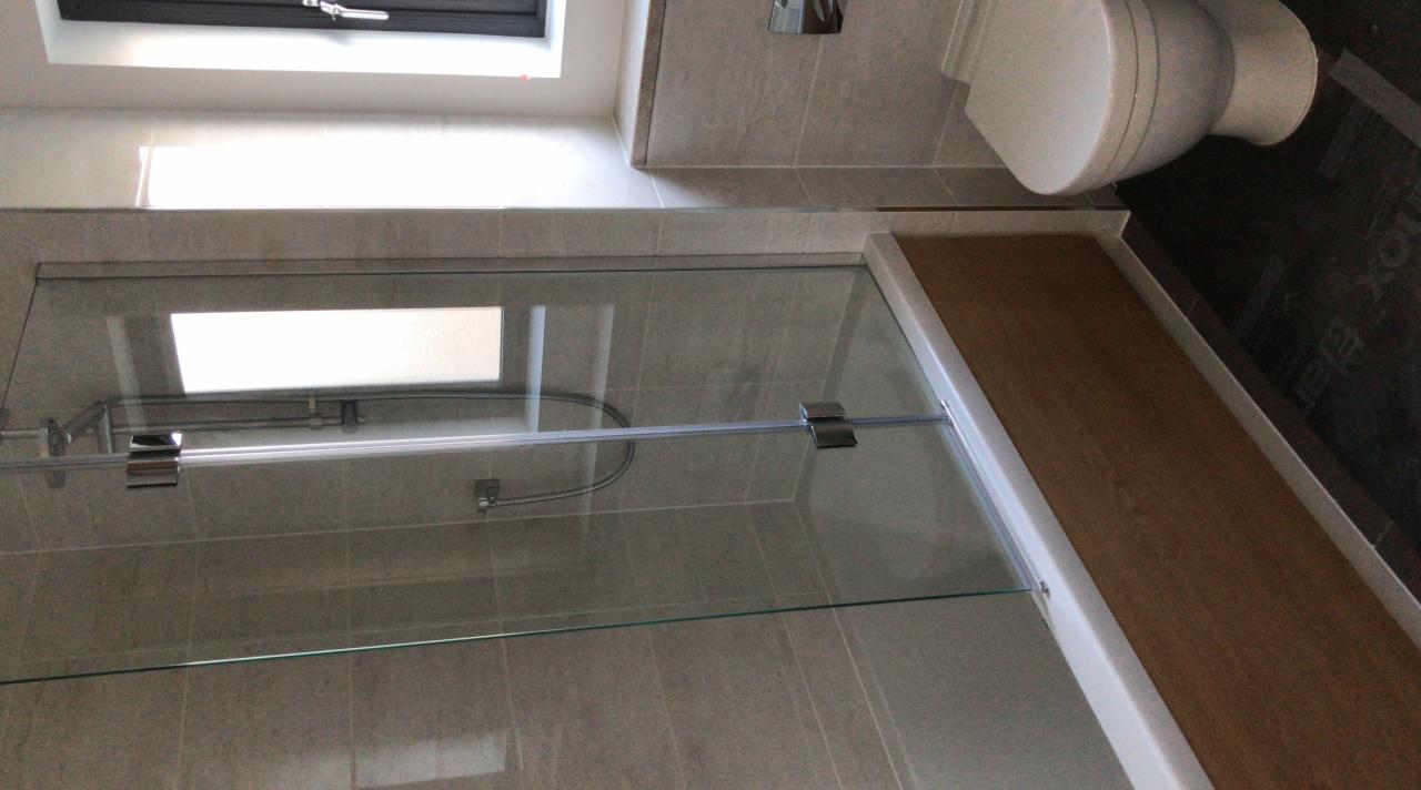 Nowoczesna kabina prysznicowa z szklanymi drzwiami, widoczny fragment toalety i okno dachowe, szare płytki na ścianach, drewnopodobny brodzik.