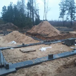 Wylane fundamenty pod budowę domu z wystającymi prętami zbrojeniowymi, otoczone piaskiem i stertami ziemi na tle lasu.