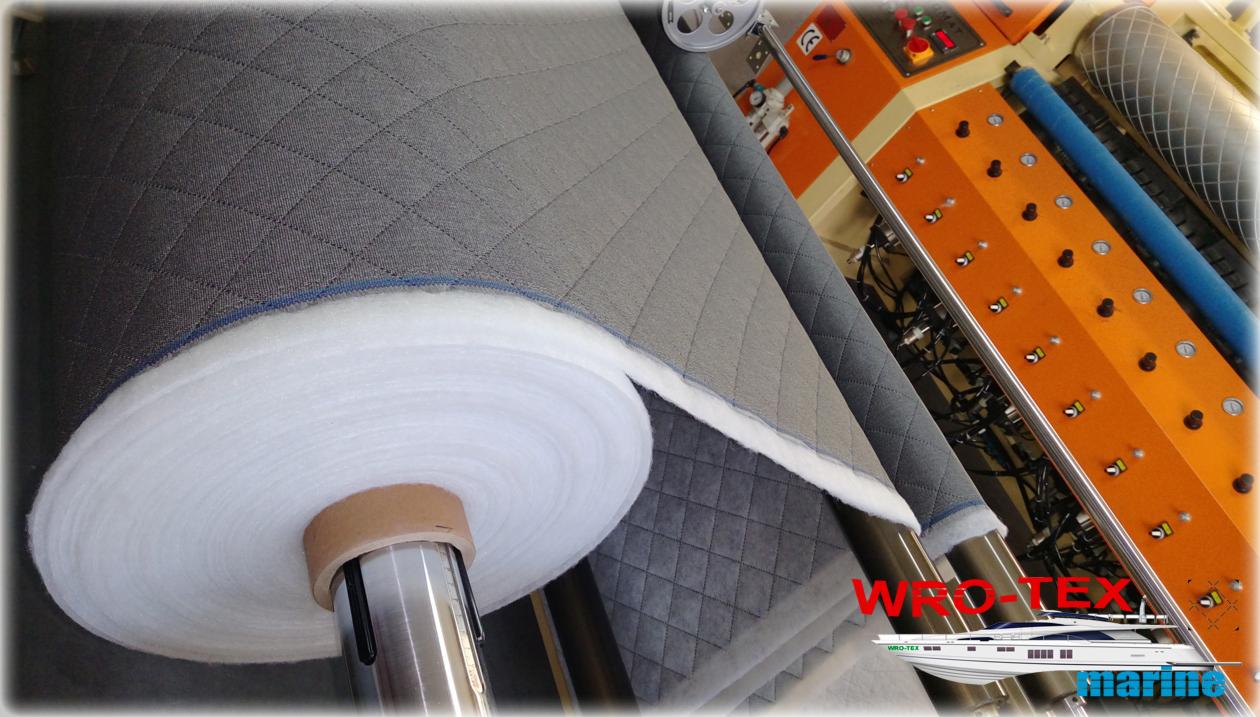 Rolka pikowanej szarej tkaniny z białym wypełnieniem na maszynie szwalniczej z logo firmy WRO-TEX marine w prawym dolnym rogu.