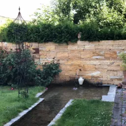 Ogród z długim, wąskim zbiornikiem wodnym wyłożonym kamieniem, kamiennym murem w tle zwieńczonym tujami, z których wystaje kamienna rzeźba głowy.