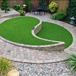 Ogród z owalnym trawnikiem z syntetycznej trawy oddzielonym niskim murem z kamienia, otoczony ścieżką z kostki brukowej, z elementami dekoracyjnymi i roślinnością ozdobną.