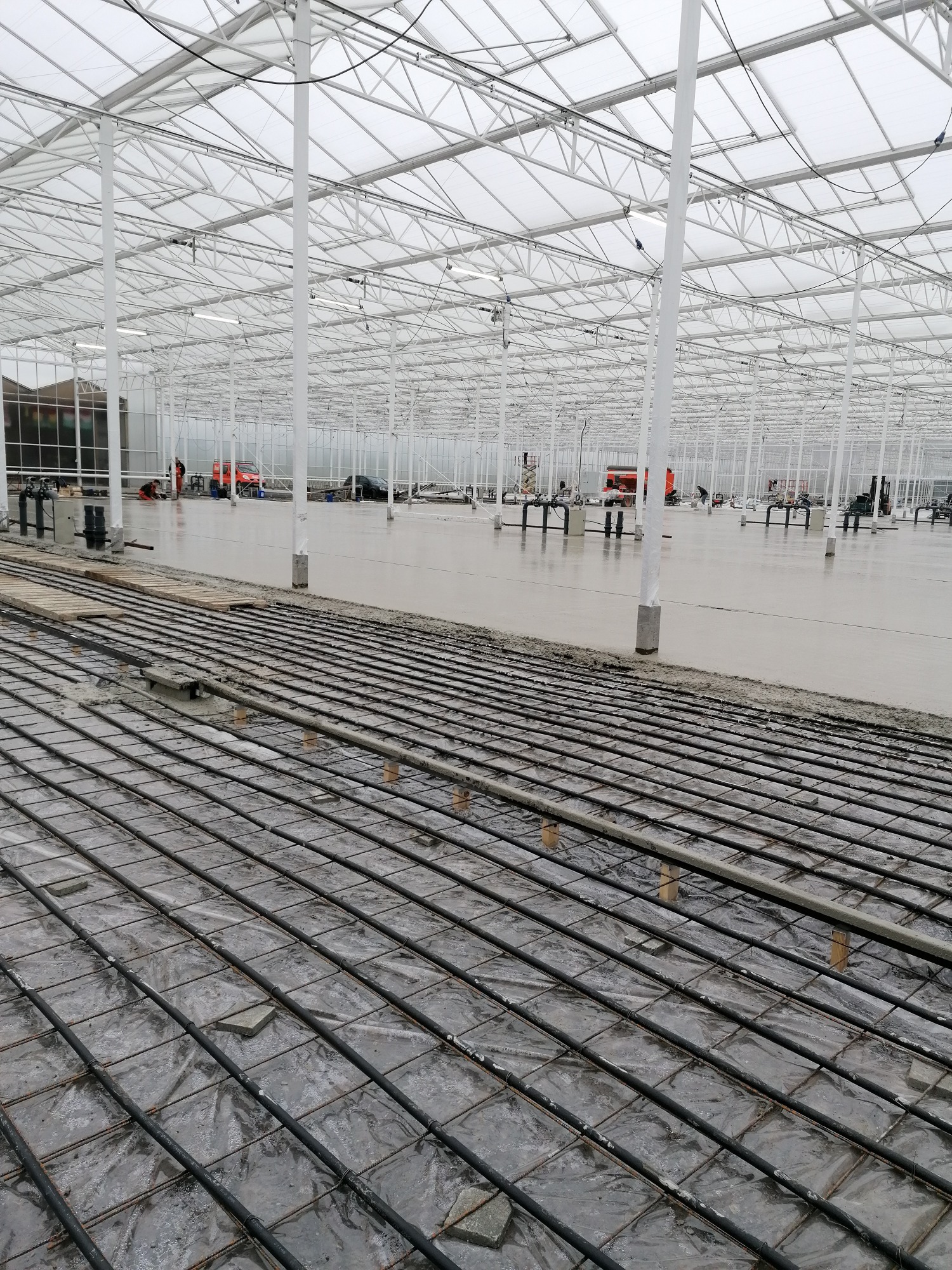 Wnętrze hali w trakcie budowy posadzki: widoczna siatka zbrojeniowa pokryta folią oraz wylewka w tle, z pracownikami i maszynami w oddali.