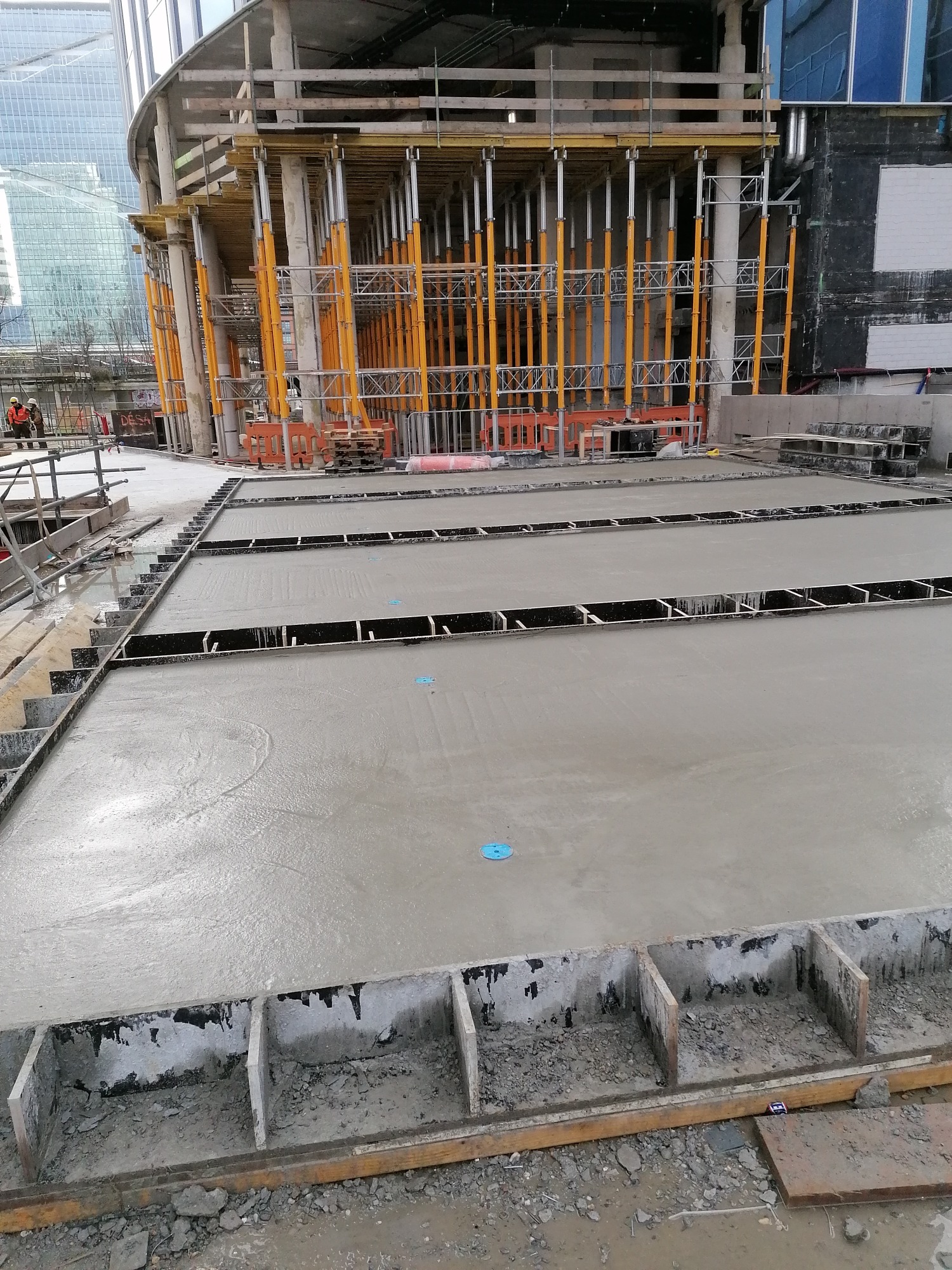 Wylewanie betonu na placu budowy z widocznymi szalunkami i wzmocnieniami, w tle konstrukcja budynku w trakcie wznoszenia z żółtymi podporami i nowoczesne wieżowce.