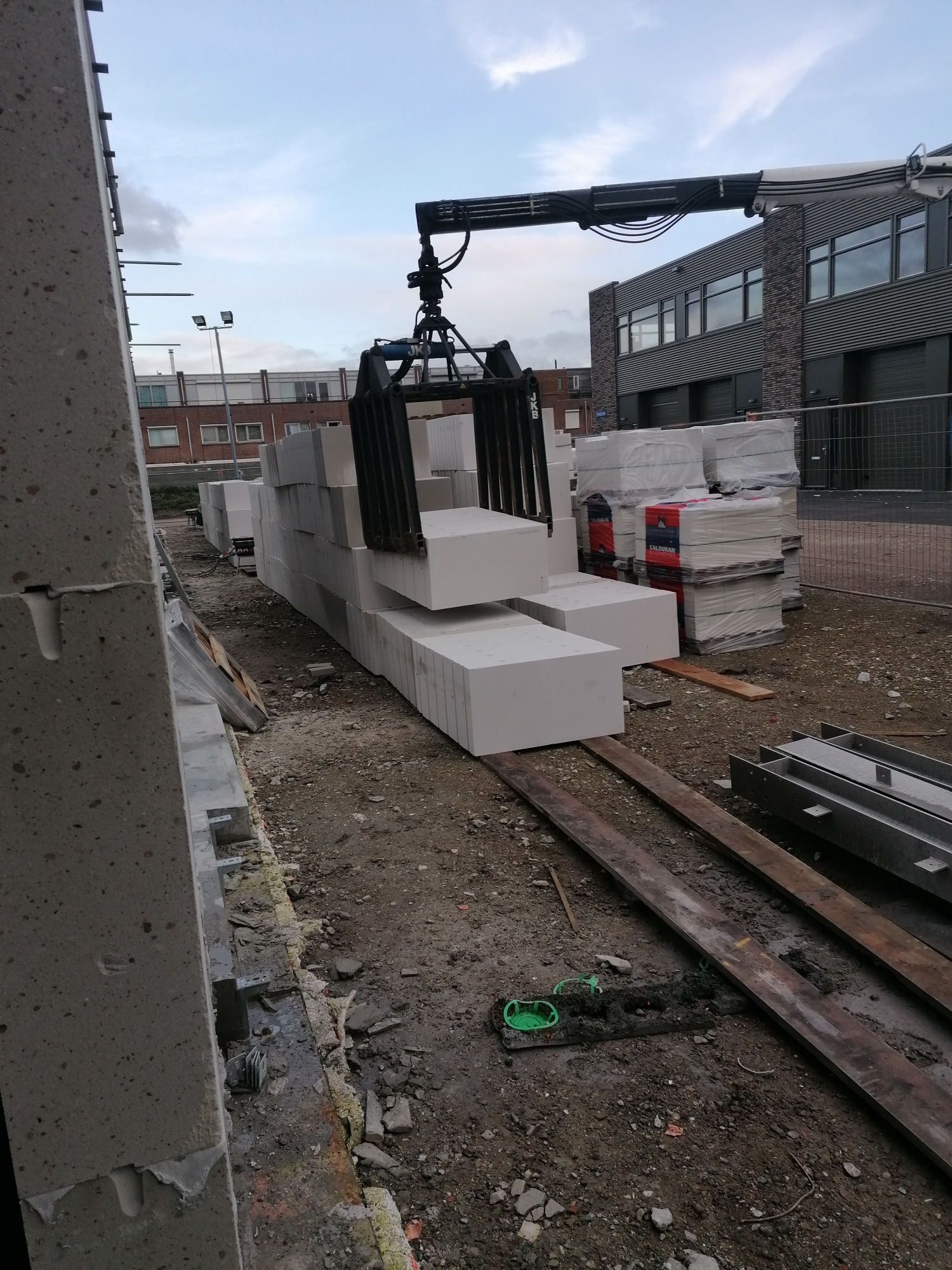 Dostawa bloczków z betonu komórkowego na plac budowy za pomocą dźwigu HDS; widoczne bloczki w uchwycie dźwigu, palety z bloczkami oraz fragment ściany w trakcie wznoszenia.