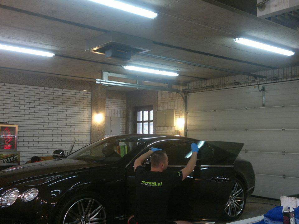 Pracownik w niebieskich rękawicach aplikuje folię na tylną szybę czarnego Bentley'a w garażu z ceglanymi ścianami i oświetleniem jarzeniowym.