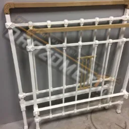 Biała, metalowa rama łóżka z dekoracyjnymi elementami, po renowacji, z widocznymi fragmentami oryginalnego, złotego koloru.