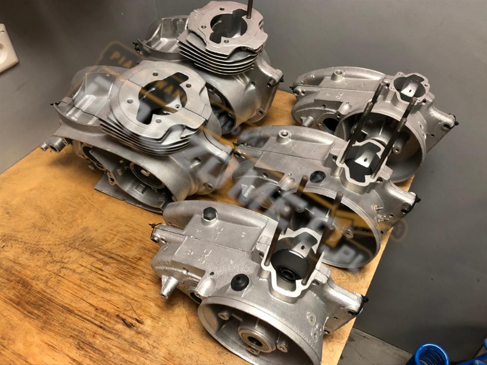 Silnik motocyklowy, aluminiowe odlewy po obróbce, leżące na drewnianym blacie warsztatowym, widoczne cylindry z żeberkami chłodzącymi i wewnętrzne elementy.