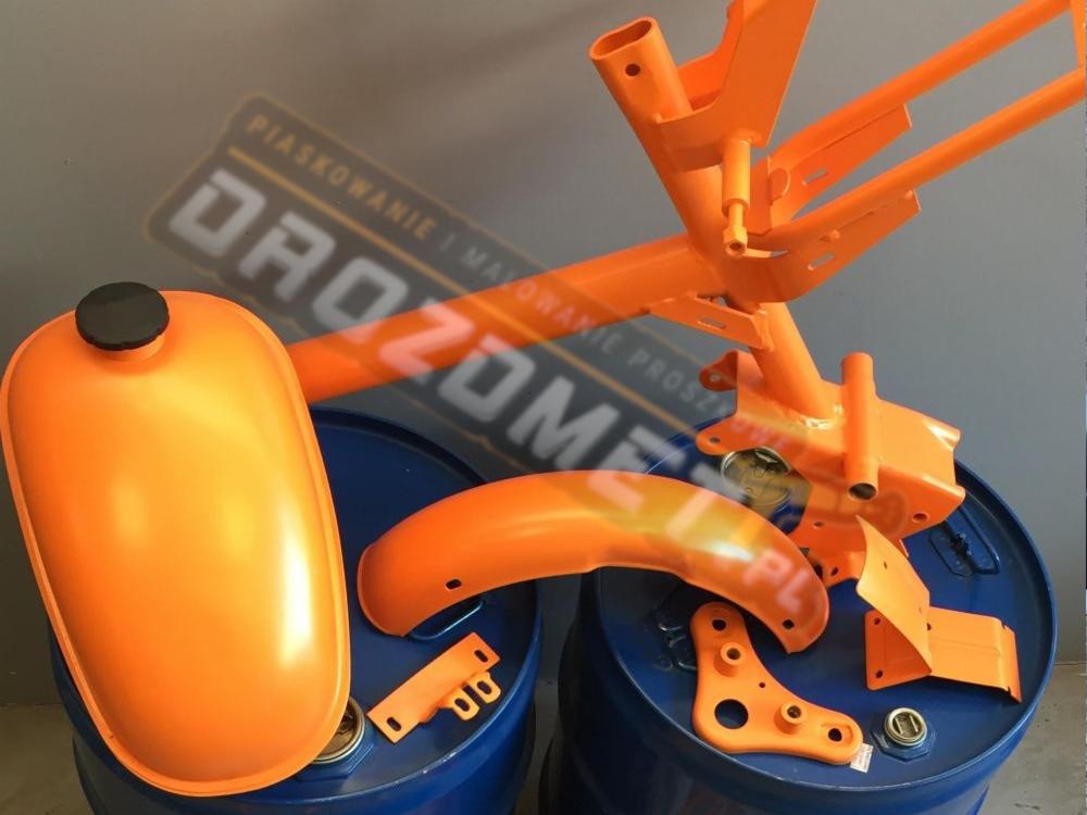 Pomarańczowe elementy motocykla: rama, błotnik, zbiornik paliwa i inne detale, po obróbce, leżące na niebieskich beczkach.