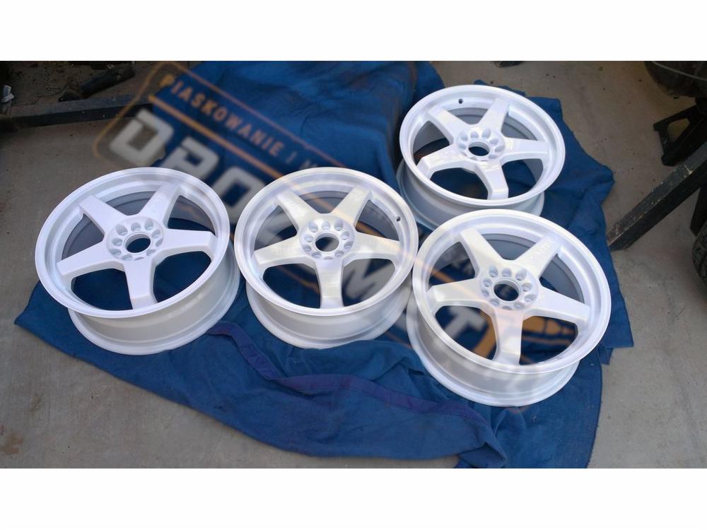 Cztery białe felgi samochodowe z pięcioramiennym wzorem, ułożone na niebieskiej szmatce, w tle widoczny fragment warsztatu.