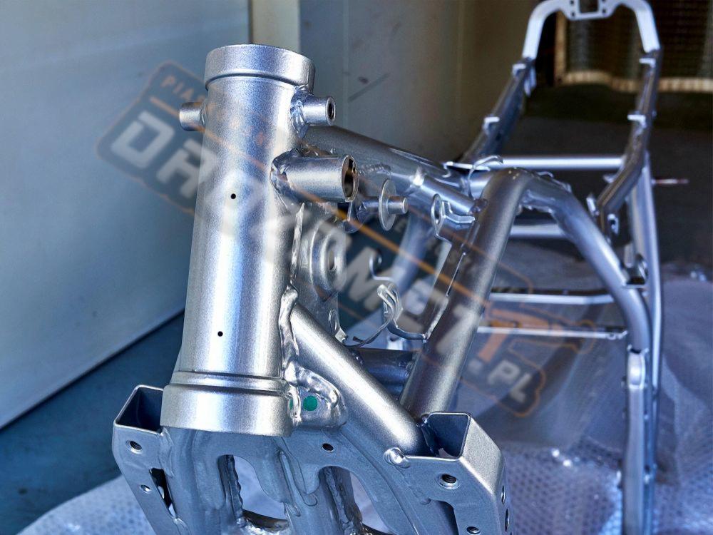 Rama motocyklowa po piaskowaniu i malowaniu proszkowym na srebrno, z widocznymi otworami montażowymi i elementami konstrukcyjnymi, w tle częściowo widoczny znak firmowy.