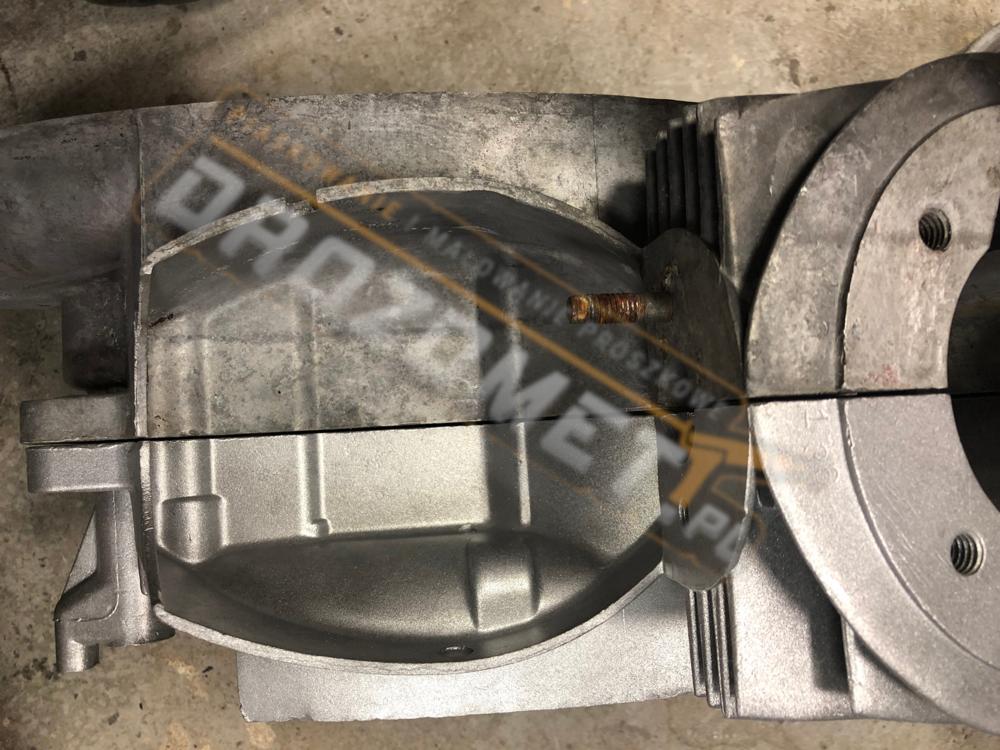 Szary, metalowy blok silnika spalinowego z widocznymi śladami obróbki, zardzewiałą śrubą i otworem na śrubę, zbliżenie.