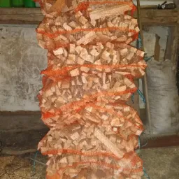 Stos worków z pociętym drewnem kominkowym w pomarańczowej siatce, ustawiony w pomieszczeniu gospodarczym o surowych ścianach, z widocznymi narzędziami i workiem w tle.