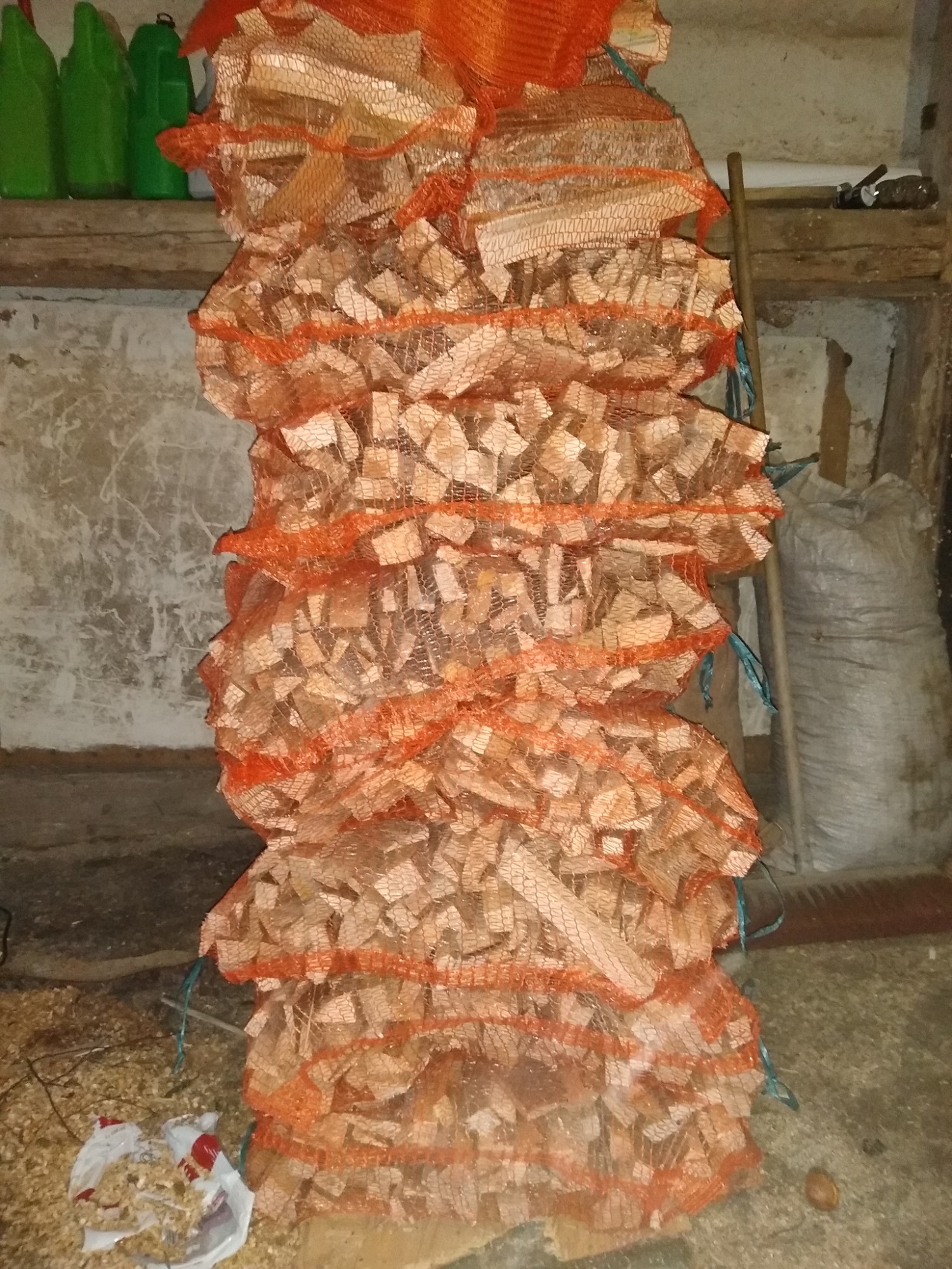 Stos worków z pociętym drewnem kominkowym w pomarańczowej siatce, ustawiony w pomieszczeniu gospodarczym o surowych ścianach, z widocznymi narzędziami i workiem w tle.