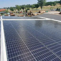 Panele słoneczne na dachu budynku przemysłowego, widoczne składowane drewno i plac manewrowy w tle.