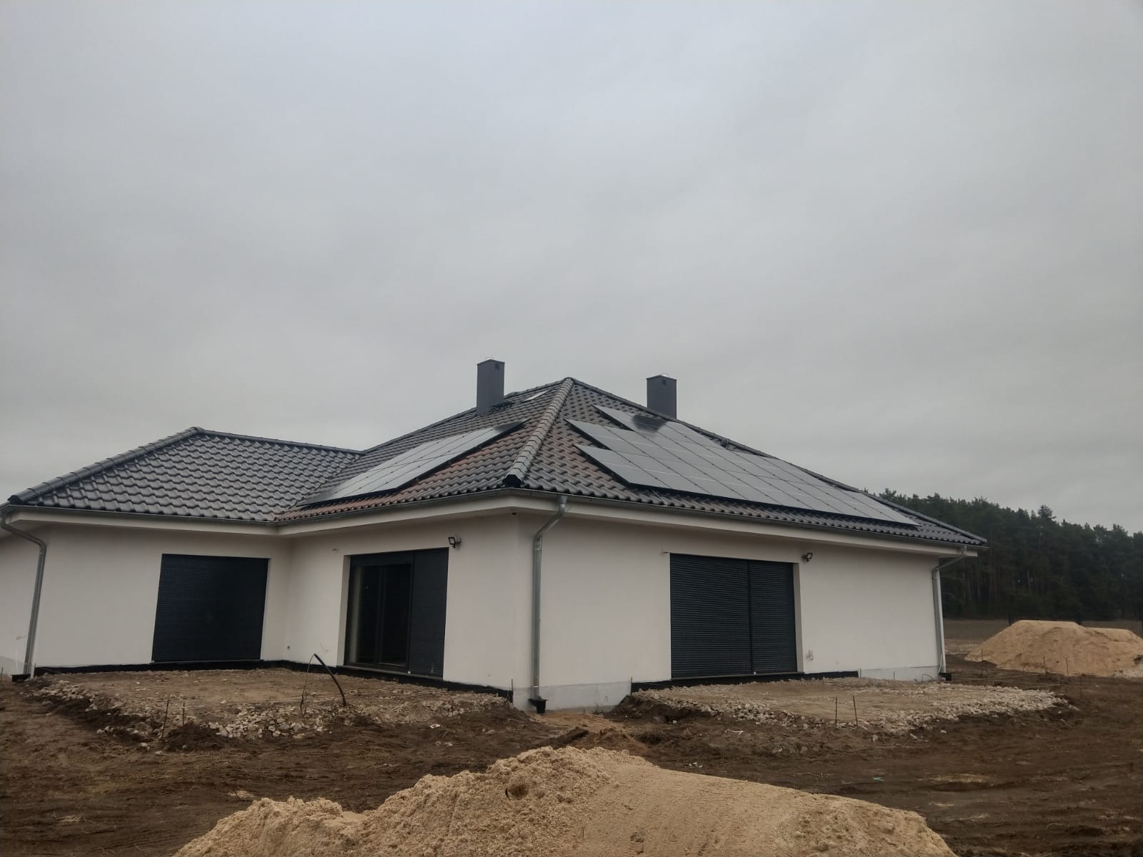 Nowoczesny dom jednorodzinny z panelami słonecznymi na ciemnoczerwonym dachu, widoczne dwa kominy, szare rolety w oknach i drzwiach balkonowych, teren wokół domu w trakcie prac ziemnych.