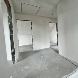 Wnętrze w stanie deweloperskim z wygładzonymi ścianami, surową podłogą i otworami drzwiowymi bez ościeżnic, widoczna klapa rewizyjna w suficie.