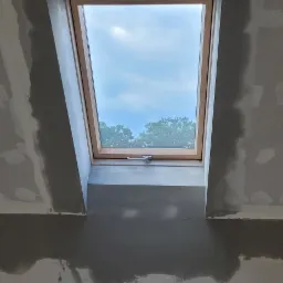 Wykończenie wnęki okna dachowego: ściany i sufit pokryte świeżo nałożoną, mokrą warstwą szarej masy szpachlowej, widoczne fragmenty odsłoniętej płyty karton-gips z miejscowymi łatami, przez okno...