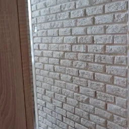 Ściana wykończona białymi płytkami imitującymi cegłę, widok z boku, fragment drewnianej ościeżnicy po lewej stronie.