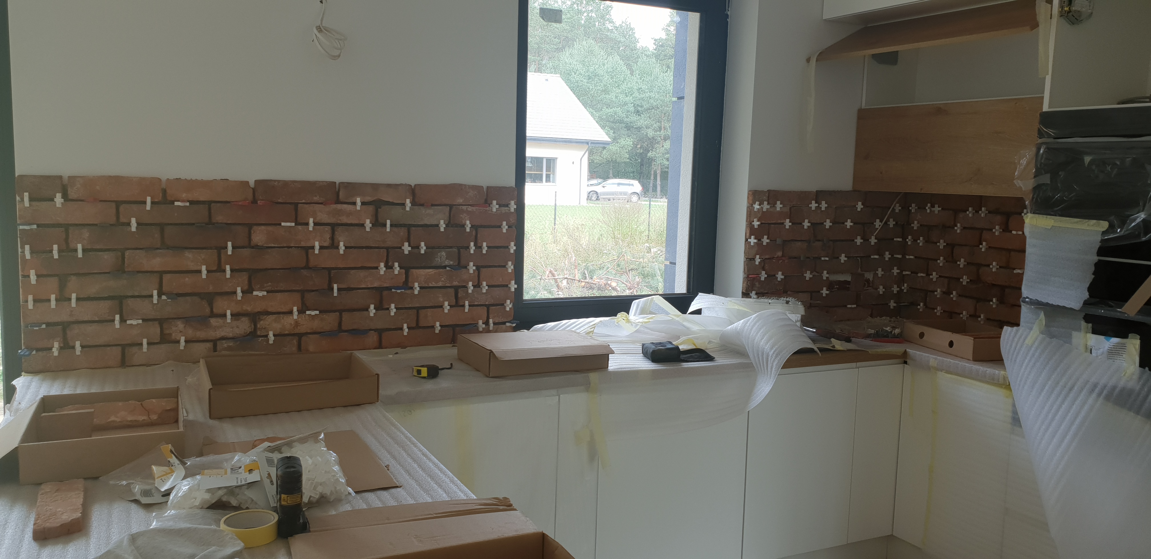 Kładzenie cegiełek dekoracyjnych na ścianie w kuchni, widoczne białe krzyżyki dystansowe, kartony, taśma miernicza i folia ochronna na blacie i szafkach, okno z widokiem na dom i zieleń.