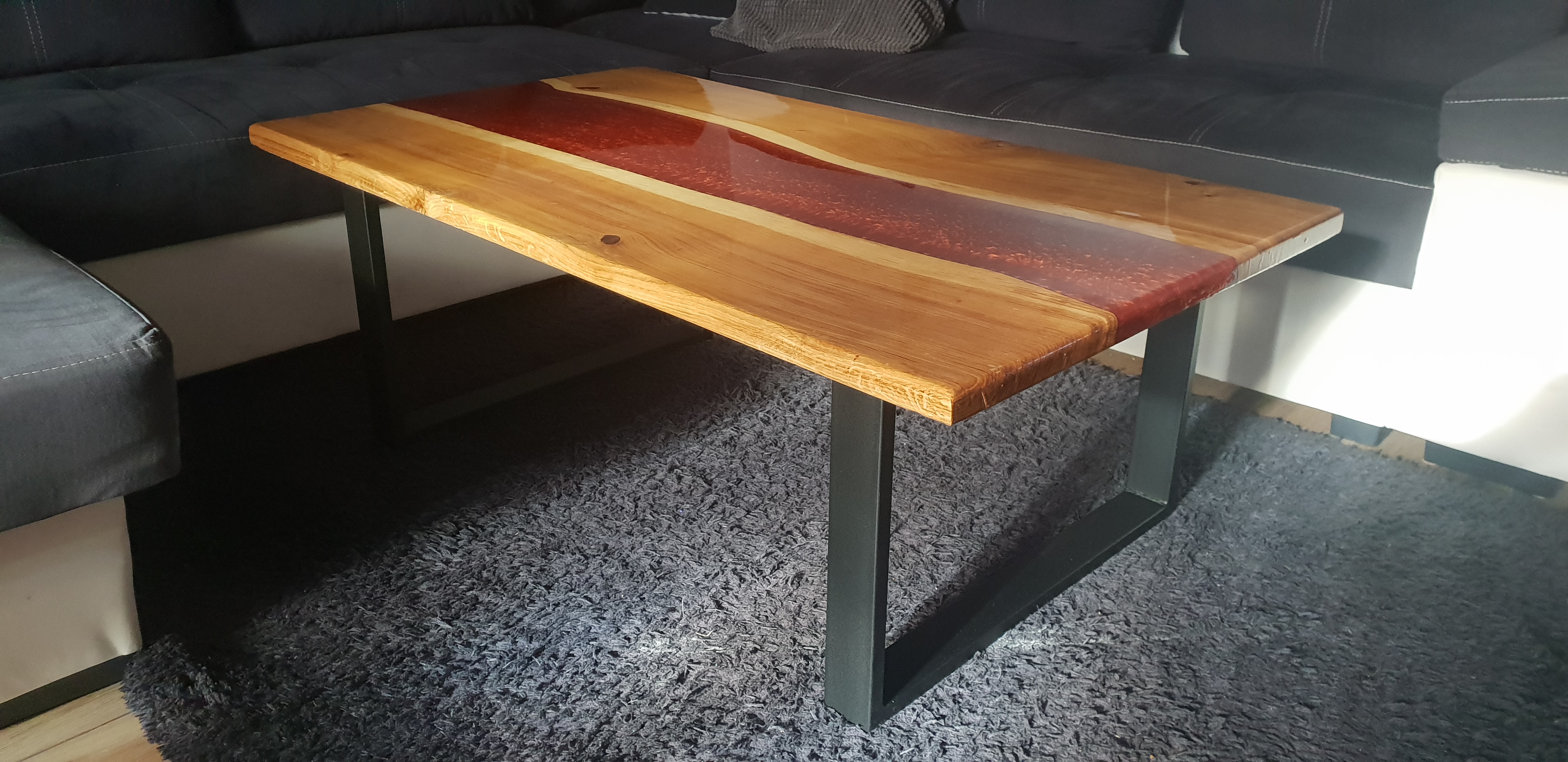 Stolik kawowy z drewna z żywicą epoksydową w odcieniach czerwieni, wsparty na metalowych nogach, ustawiony na szarym dywanie w salonie.