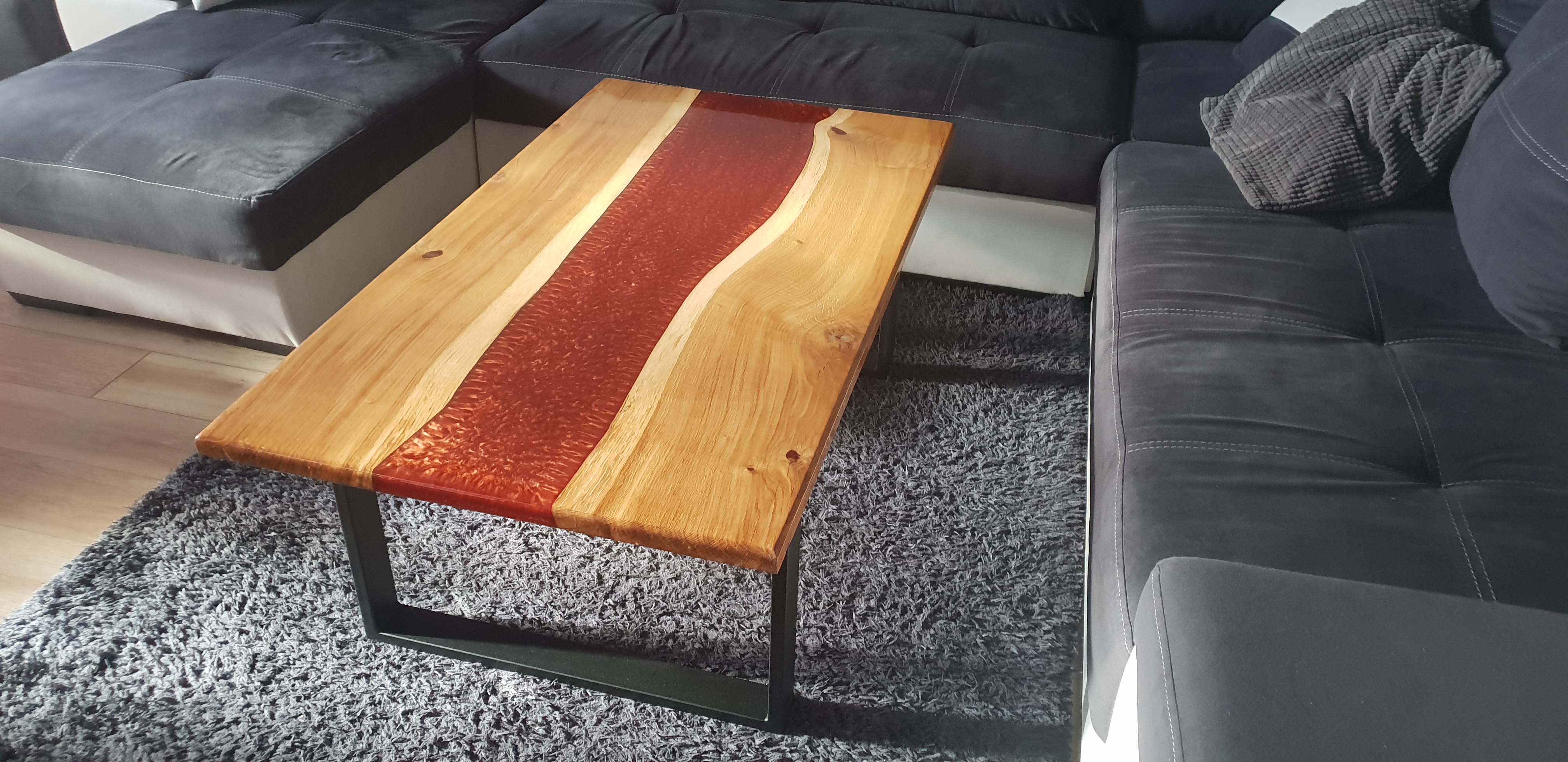 Stolik kawowy z blatem z jasnego drewna, z żywicą epoksydową w kolorze czerwonym, imitującą rzekę, na tle szarego dywanu i ciemnej sofy.