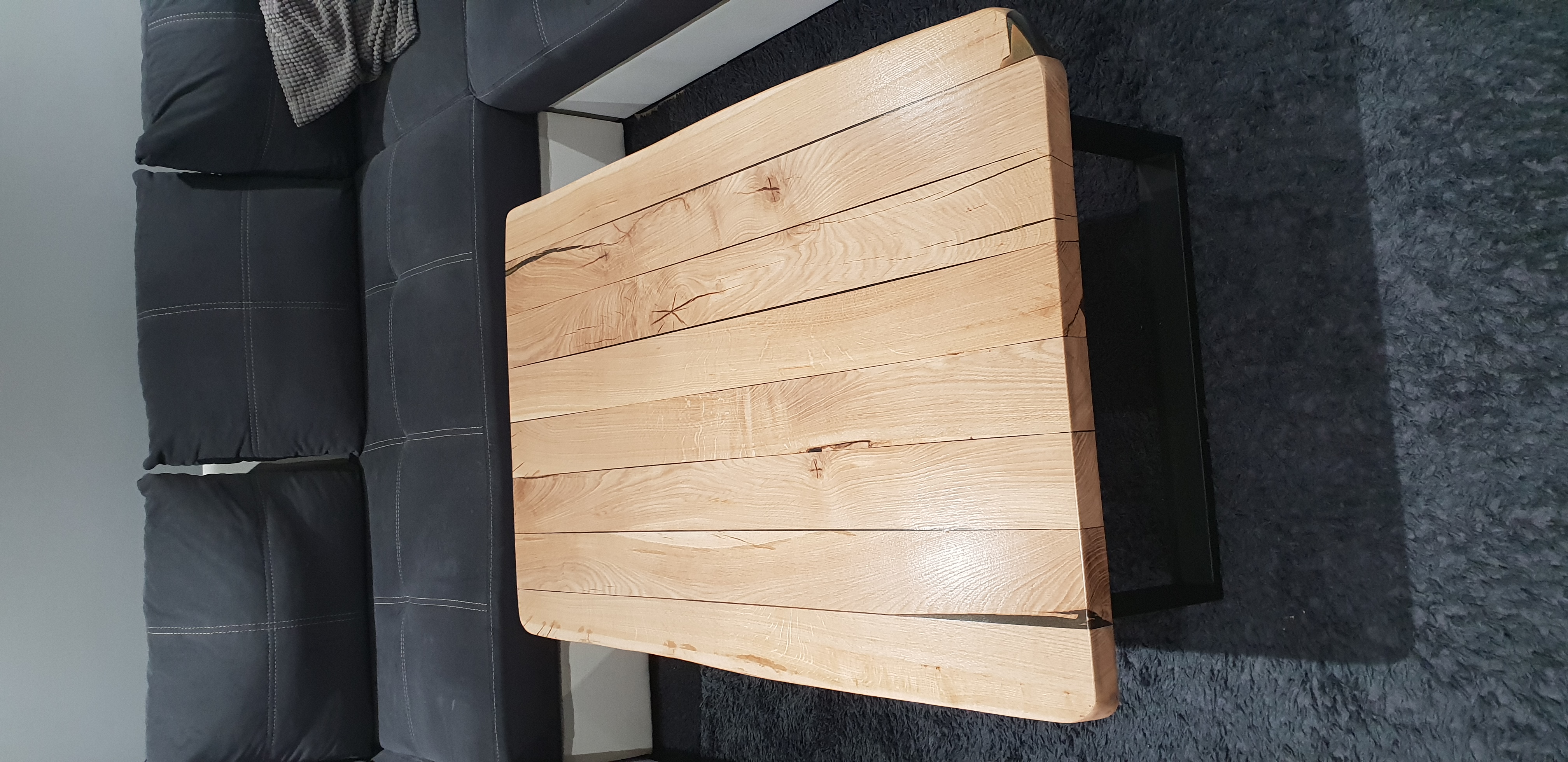 Stolik kawowy z jasnego drewna, złożony z kilku desek z widocznymi naturalnymi pęknięciami, na czarnych metalowych nogach, obok szarej kanapy i dywanu.