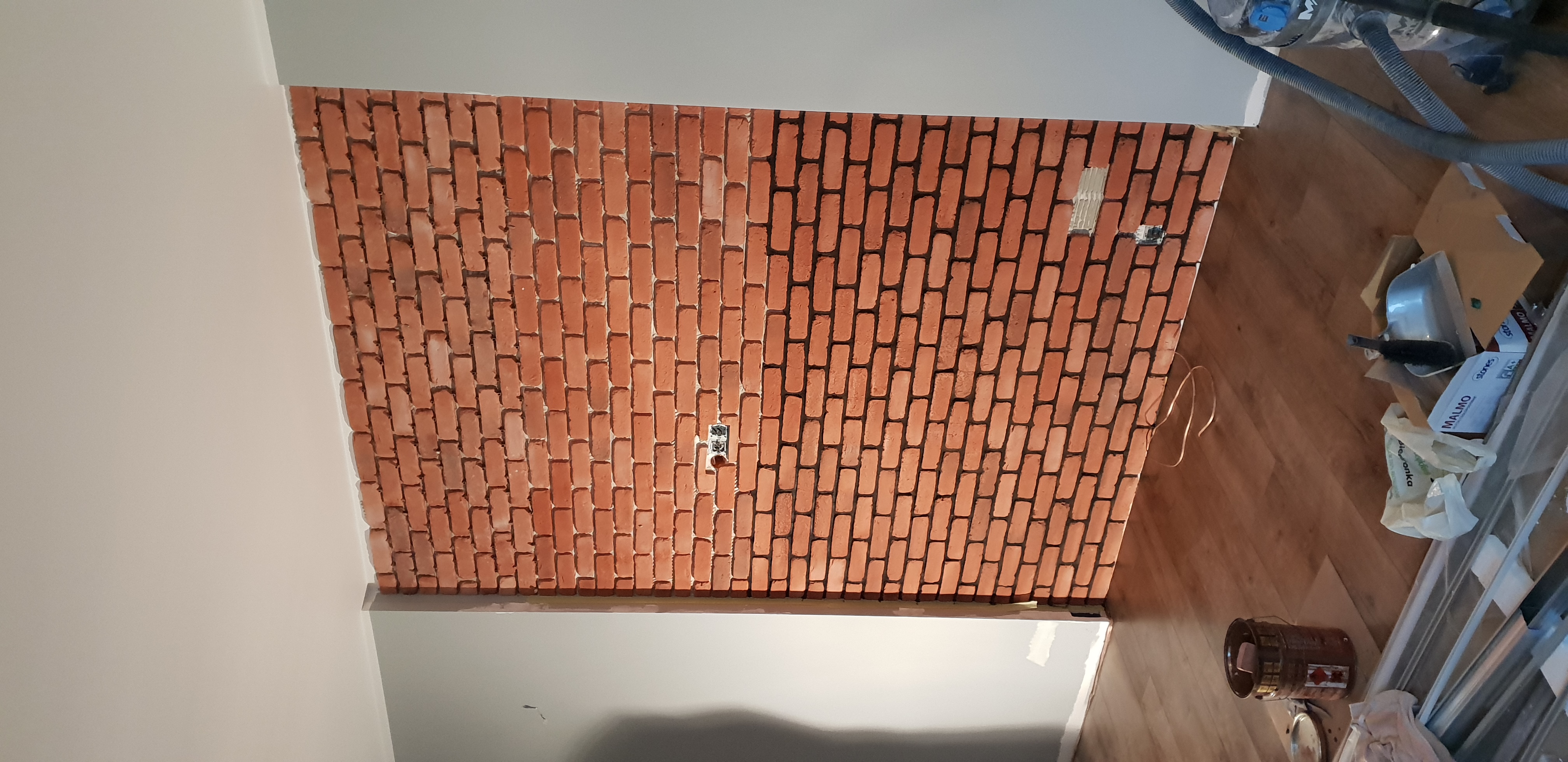 Ściana z cegieł klinkierowych wewnątrz pomieszczenia, z widocznymi puszkami elektrycznymi i fragmentem sufitu oraz podłogi z narzędziami i akcesoriami malarskimi.