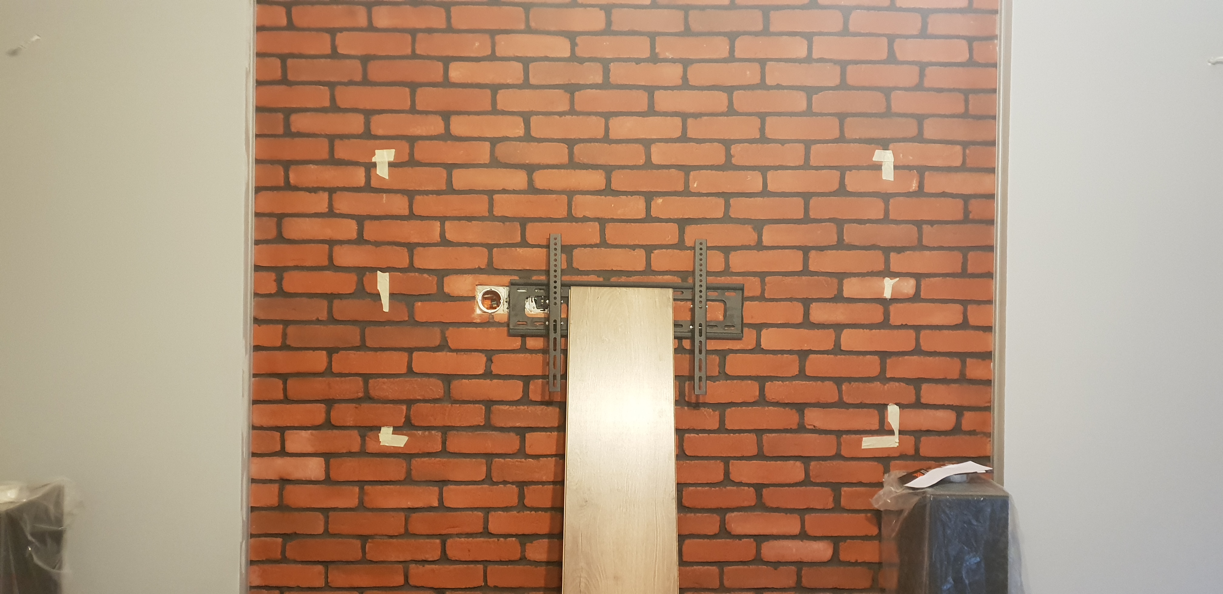 Montaż płyty karton gips na ścianie z tapetą imitującą czerwoną cegłę, widoczny uchwyt do telewizora, puszka elektryczna i fragment płyty oraz taśmy maskujące.