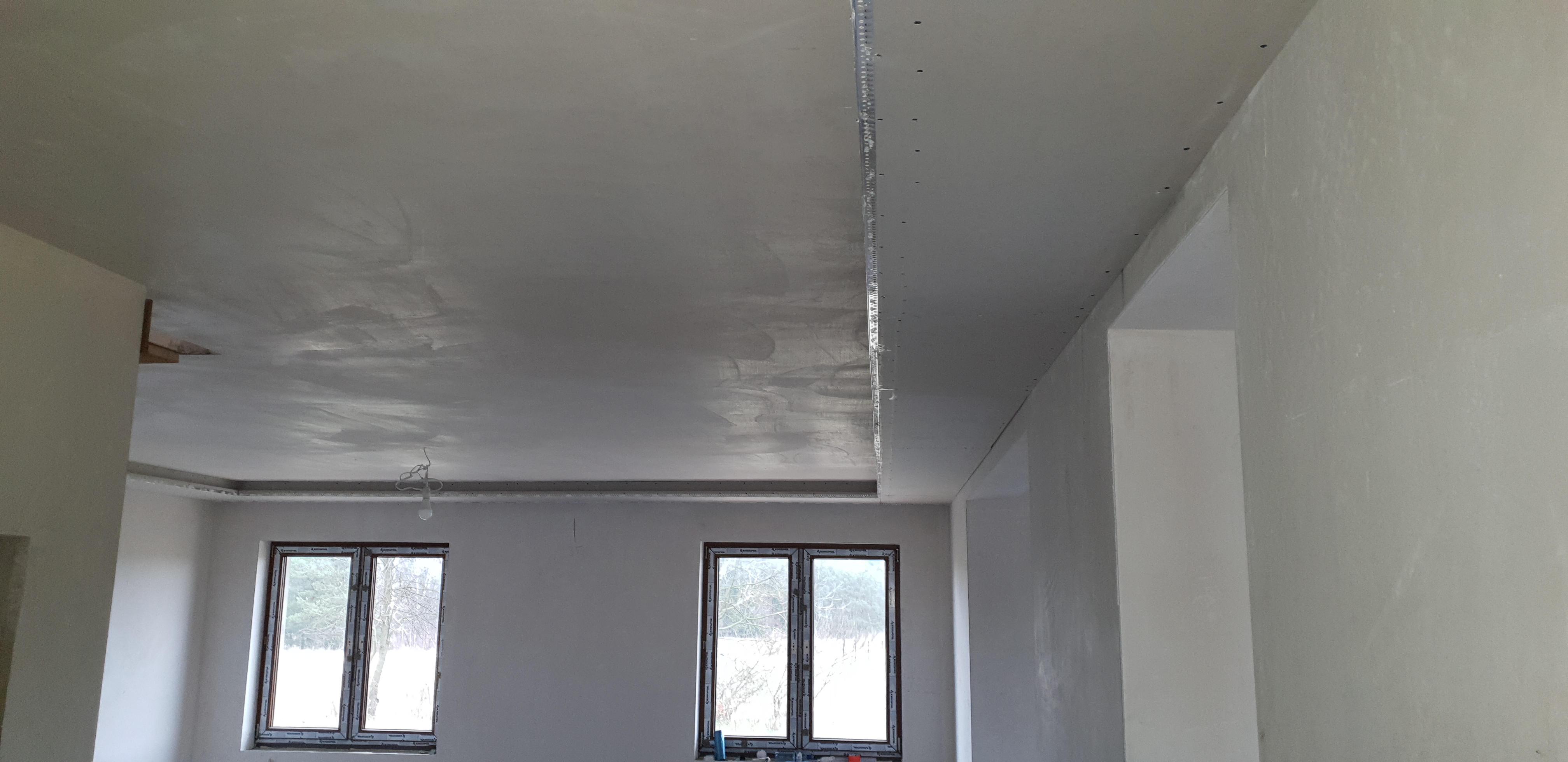 Świeżo położona gładź na suficie z widocznymi profilami do montażu oświetlenia, surowe ściany w tle, dwa okna z widokiem na zimowy krajobraz.