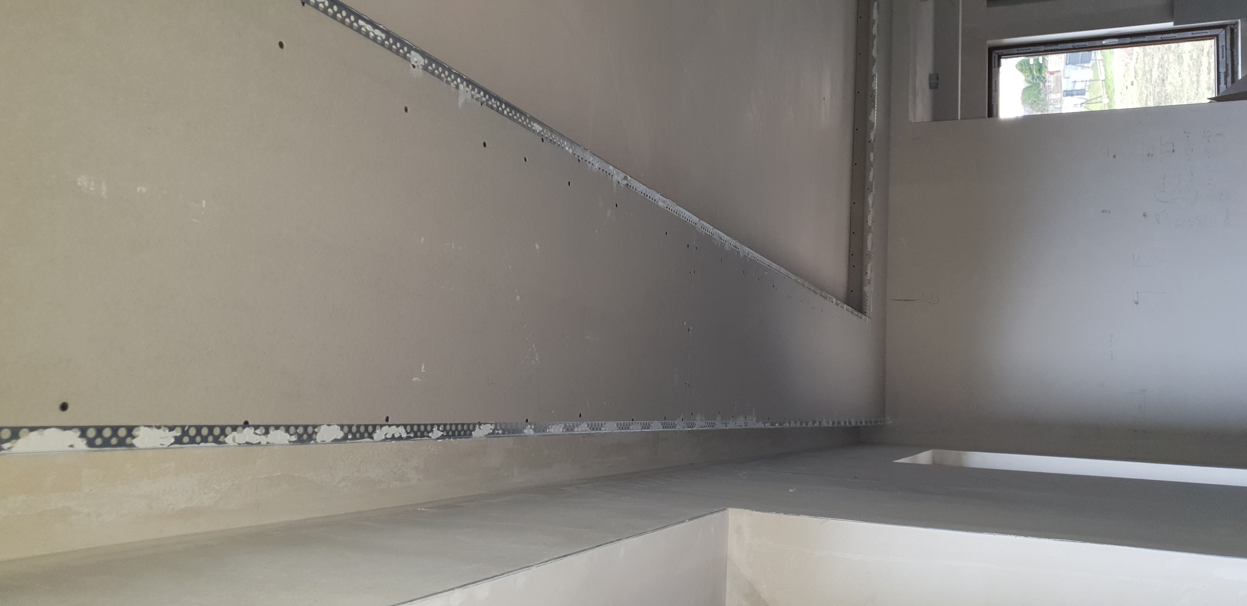 Surowe wnętrze w trakcie prac wykończeniowych: ściany z płyt kartonowo-gipsowych z widocznymi listwami perforowanymi, przygotowane do dalszych etapów prac, zarys okna w tle.