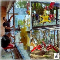 Montaż dużego okna z użyciem specjalistycznego podnośnika próżniowego, widoczny pracownik regulujący położenie szyby, w tle budynek w stanie surowym i drzewa.