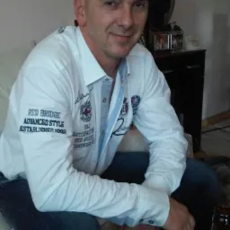 Mężczyzna w białej koszuli siedzi na kanapie w pomieszczeniu. W tle widać wentylator i dekoracje na komodzie, butelka piwa w prawym dolnym rogu.