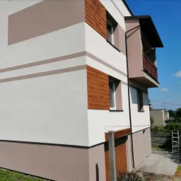 Dwupiętrowy dom z jasną elewacją, brązowymi akcentami w postaci pasów i paneli imitujących drewno, balkon z drewnianą balustradą, widoczne elementy instalacji odgromowej.