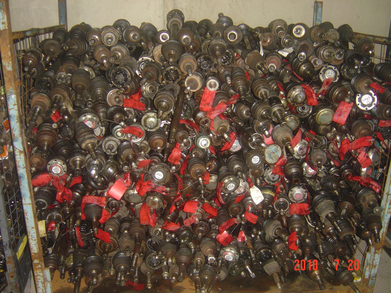 Metalowa klatka wypełniona po brzegi używanymi półosiami samochodowymi z czerwonymi etykietami, widoczne ślady użytkowania i różnice w modelach.