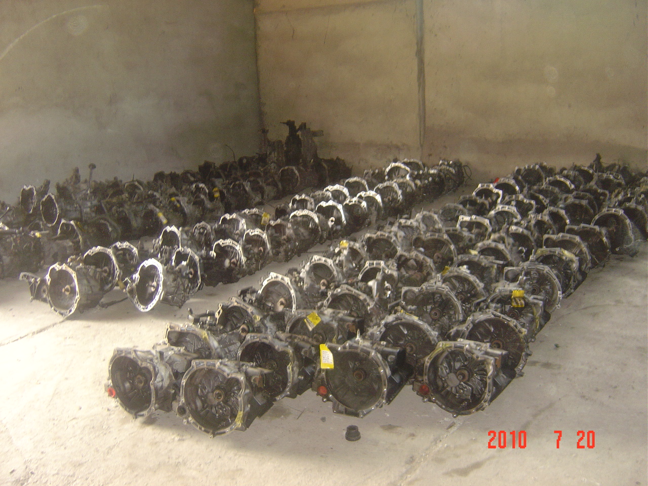 Magazyn używanych skrzyń biegów samochodowych, ułożonych rzędami na betonowej podłodze, widoczne żółte etykiety cenowe na niektórych egzemplarzach, data na zdjęciu 2010-07-20.