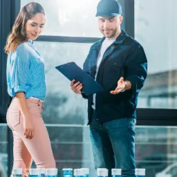 Mężczyzna w roboczym stroju i czapce wręcza kobiecie dokumenty na tle butli z wodą, jasne światło z okna.