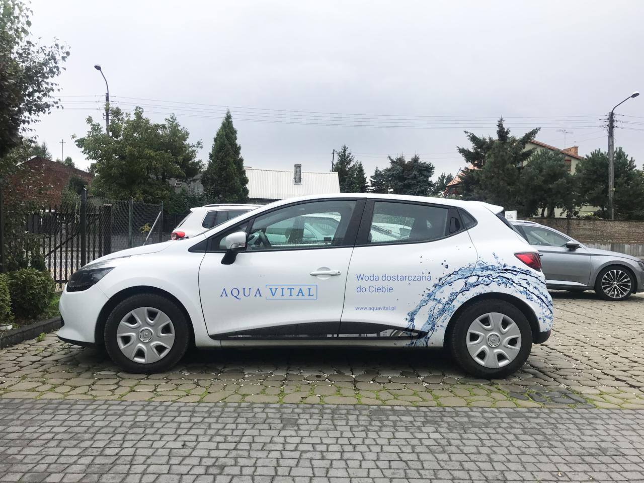 Biały samochód marki Renault Clio z logo 'Aqua Vital' i hasłem 'Woda dostarczana do Ciebie' na boku, zaparkowany na brukowanym parkingu w pochmurny dzień.
