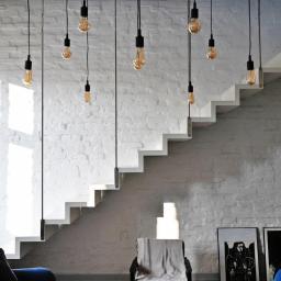 Pracownia Snycerska u Pawła - Nowoczesne schody betonowe przy białej ceglanej ścianie, oświetlone wiszącymi żarówkami na czarnych kablach, widok z salonu z niebieską kanapą.