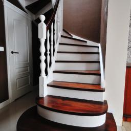 Pracownia Snycerska u Pawła - Kręte schody z ciemnego drewna i białymi podstopnicami, biała balustrada z drewnianą poręczą, widok z dołu, w tle drzwi.