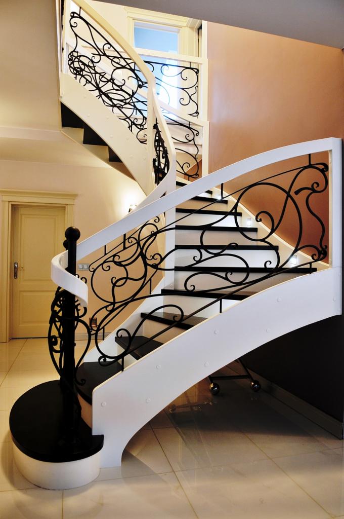Kręte schody z czarnymi stopniami i białymi bokami, ozdobione ażurową, czarną balustradą ze stylizowanymi motywami roślinnymi.