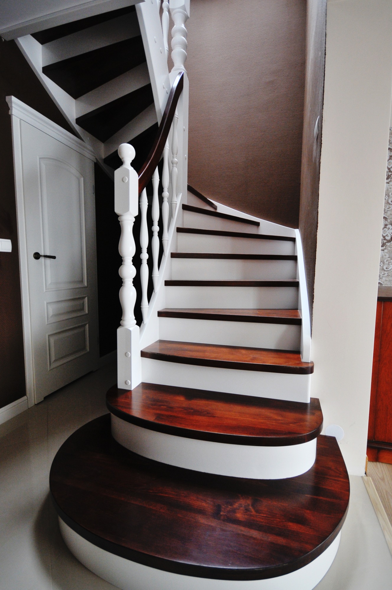 Kręte schody z ciemnego drewna i białymi podstopnicami, biała balustrada z drewnianą poręczą, widok z dołu, w tle drzwi.