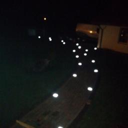 FHU Dariusz Bojaś - Oświetlona ścieżka w ogrodzie nocą, z lampami punktowymi wzdłuż drewnianego chodnika prowadzącego do domu.