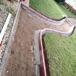 FHU Dariusz Bojaś - Układanie obrzeży trawnikowych z czerwonej kostki, widoczny młotek i zaprawa cementowa, trawa po prawej stronie.