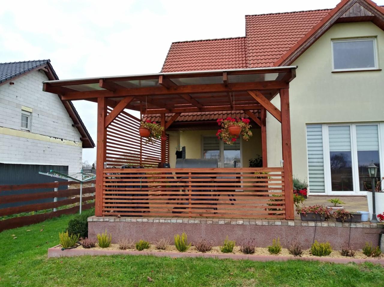 Drewniana pergola z przezroczystym zadaszeniem i poziomymi listwami, z wiszącymi donicami z czerwonymi kwiatami, przylegająca do jasnego domu z oknami PCV, widok z ogrodu.