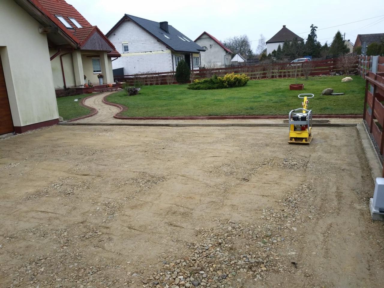 Przygotowanie podłoża pod kostkę brukową z użyciem zagęszczarki w przestrzeni przed domem jednorodzinnym, widoczny fragment chodnika i trawnika.