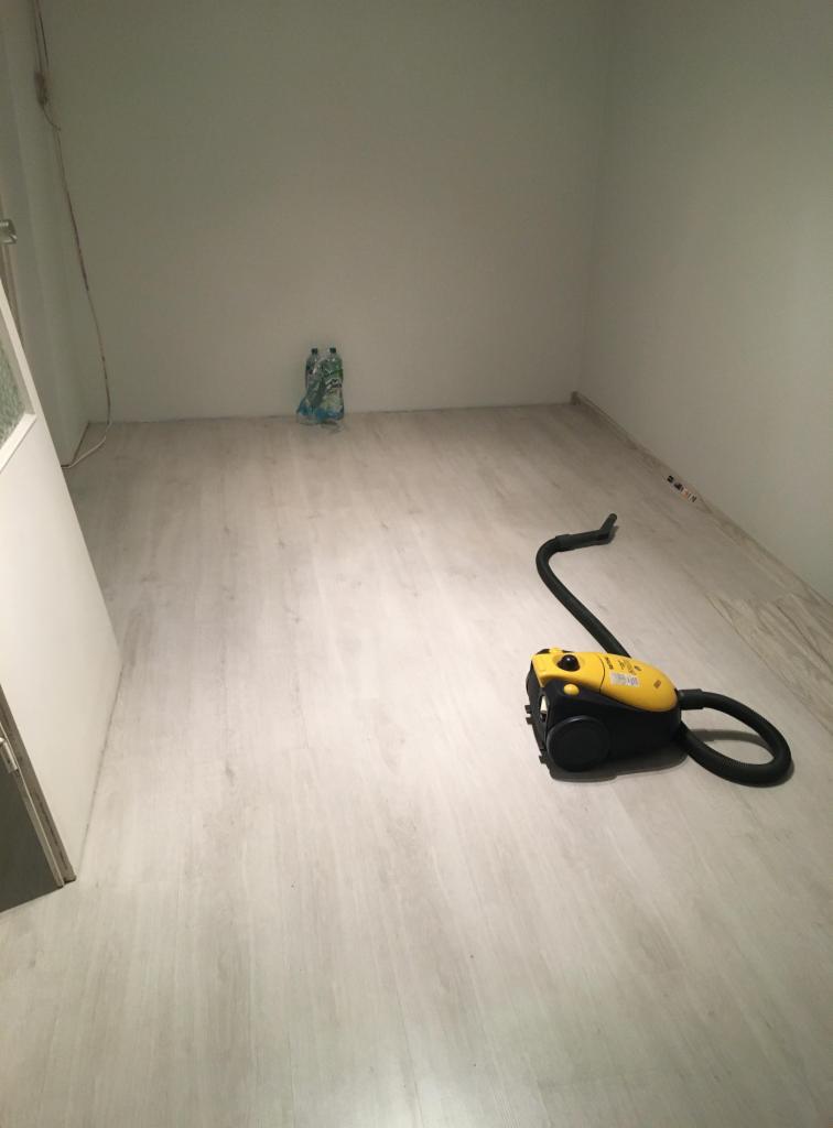 Jasne panele podłogowe w pustym pokoju, żółto-czarny odkurzacz na podłodze, butelki z wodą pod ścianą.